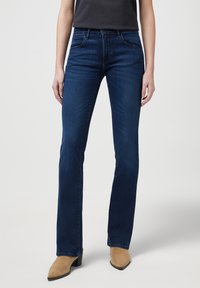 Jeans en denim évasés de couleur bleu foncé avec une texture lisse, dotés de poches avant et d'une taille moyenne, associés à des bottines beige à bout pointu.