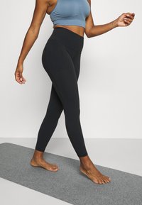 Fekete, magas derekú leggings, letisztult dizájnnal és sima textúrával, világoskék crop top-pal párosítva, egy sétáló pózban, szürke szőnyegen bemutatva.