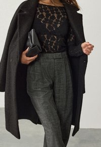 Manteau en laine noire porté sur un haut en dentelle noire, associé à un pantalon gris texturé. La tenue est complétée par une pochette noire, mettant en valeur les matériaux contrastés.