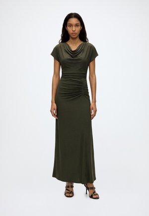 Mujer de pie con un vestido largo color verde oliva con detalles fruncidos y sandalias negras de tacón alto sobre un fondo blanco.