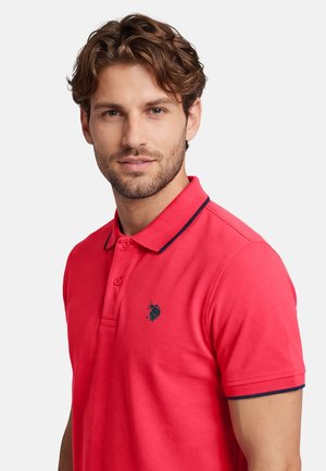 Young man with brown hair wearing a red polo shirt with navy trim and small embroidered logo, looking slightly toward the camera.