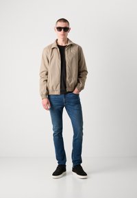 Beige Bomberjacke mit Reißverschluss, schwarzes T-Shirt, blaue Jeans und schwarze Sneakers. Zubehör umfasst dunkle Sonnenbrillen.