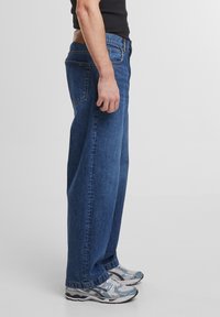 Jean en denim bleu à coupe ample avec une conception à jambe droite, cinq poches et une étiquette en cuir beige à l'arrière de la taille. Porté avec des chaussures de sport grises.