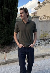 Mladý muž v tmavozelenej polokošeli a tmavých džínsoch stojí na ceste pri vysokom strome a kamennej stene s domami v pozadí.