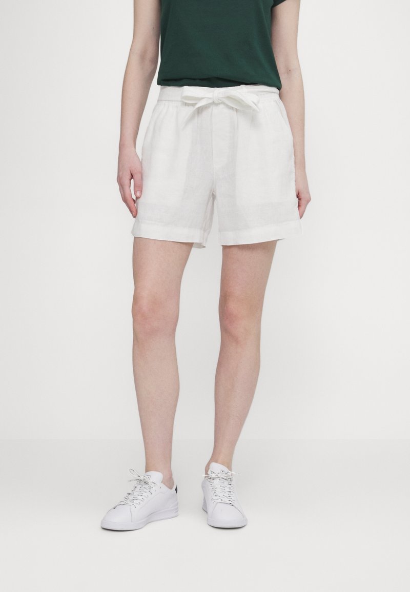 Personne portant un short blanc taille haute avec une ceinture nouée, un haut vert foncé et des baskets blanches, debout devant un fond uni.