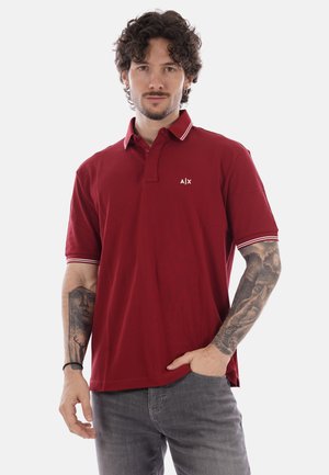 Uomo con i capelli ricci che indossa una polo rossa con rifiniture bianche e jeans grigi, mostrando le braccia tatuate, in piedi davanti a uno sfondo semplice.