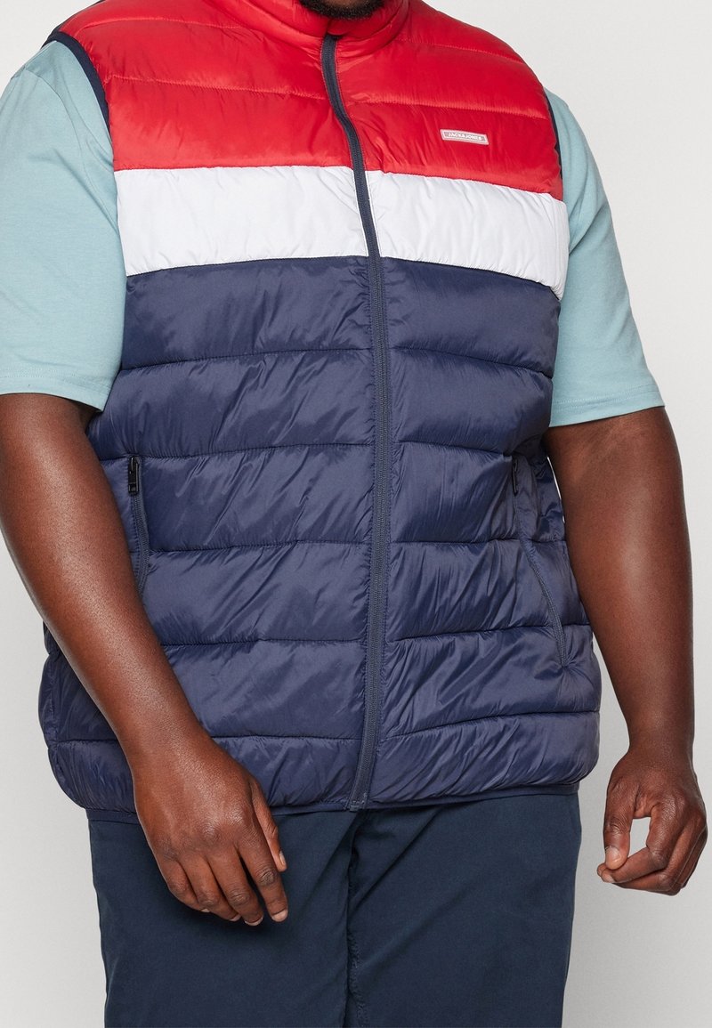 Chaleco acolchado multicolor con un diseño de paneles en rojo, blanco y azul marino. El material parece suave y ligero, con bolsillos laterales con cremallera.
