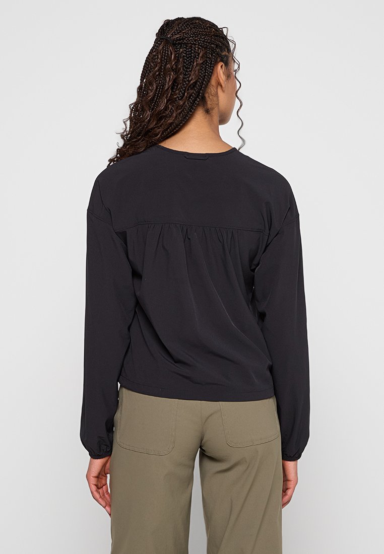 Frau mit geflochtenem Haar, die ein lockeres schwarzes Langarmoberteil und olivgrüne Hosen trägt, steht mit dem Rücken zu einer schlichten hellen Hintergrund.