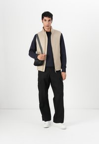 Beige gepolsterte Weste über einem marineblauen Sweatshirt, schwarze Cargo-Hosen und weiße Sneaker; hält eine schwarze Umhängetasche; einfarbiger Hintergrund.
