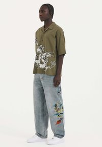 Olijfgroene shirt met korte mouwen en witte draakprint, gecombineerd met lichte spijkerbroek met kleurrijke draakborduursels op het been. Witte sneakers.