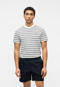 Άνδρας με κοντομάνικο πουκάμισο με λευκές και ναυτικές ρίγες και μαύρα σορτς, στέκεται μπροστά από απλό λευκό φόντο.