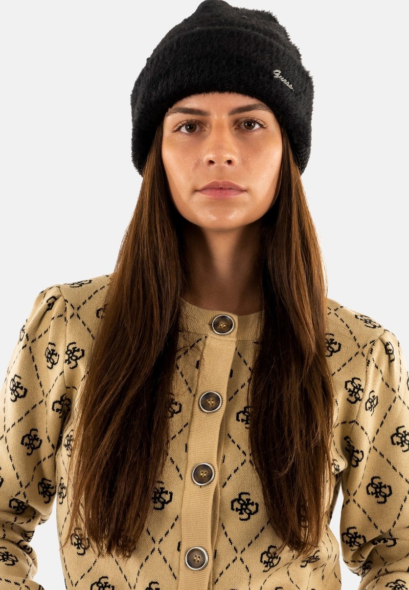 Cárdigan de punto beige con patrón floral negro y botones grandes, combinado con un gorro negro de felpa. El cabello largo y castaño enmarca el rostro.