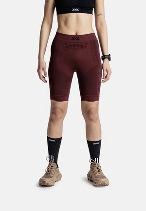 Atletisch persoon met bordeaux compressieshorts, een zwarte sport-bh, zwarte kuitkousen, beige trailrun-schoenen en een fitnesshorloge.