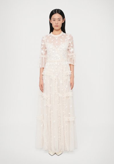 Longue robe ivoire avec superposition en dentelle florale transparente, détails volantés et manches cloches. Taille cintrée menant à une jupe fluide à étages.