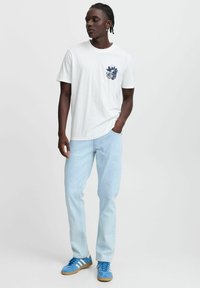 Hvit t-skjorte i bomull med et marineblått grafisk motiv på venstre bryst, kombinert med lyseblå denimjeans og blå atletikksko med hvite striper.