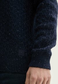 Donkerblauwe gebreide trui met een gewatteerd patroon en geribde boorden. Bevat een klein zwart label met een logo nabij de zoom.