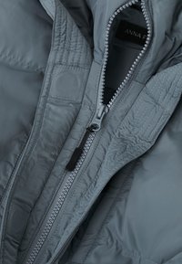 Siva puffer jakna s zatvaranjem na patentni zatvarač i crnom povlačnom trakicom, s kviltanom teksturom i glatkim, sjajnim materijalom. Postava vidljiva na ovratniku.