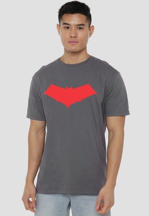 Young man wearing a gray T-shirt with a large red bat symbol on the chest, standing against a plain light background.