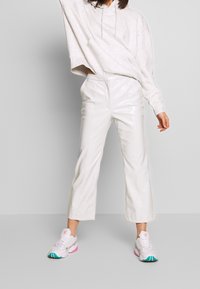 Weiße, kurz geschnittene Hosen aus Kunstleder kombiniert mit einem hellgrauen, übergroßen Hoodie. Das Model trägt weiße Sneaker mit rosa und türkisfarbenen Akzenten.