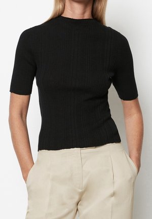 Kvinde iført en tætsiddende sort ribbet kortærmet top, kombineret med højtaljede beige bukser, hænderne afslappet i lommerne.