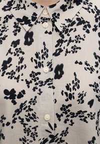 Ανοιχτό μπεζ πουκάμισο με μαύρο αφηρημένο ανθικό μοτίβο, μυτερό γιακά και τέσσερα μικρά λευκά κουμπιά μπροστά.