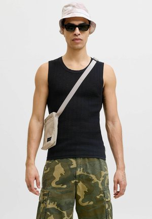 Muž nosiaci čierne tielko bez rukávov, maskáčové cargo kraťasy, klobúk typu bucket, slnečné okuliare a béžovú kabelku cez rameno na jednoliatom pozadí.