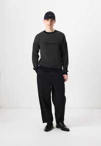 Pulover tricotat negru cu un model texturat și logo pe piept, asortat cu pantaloni negri largi și pantofi din piele negri, purtat cu o șapcă neagră.
