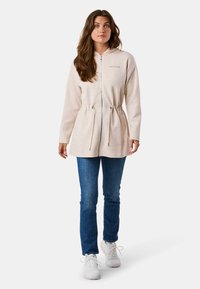 Haut à capuche zippé beige clair avec taille à cordon, manches longues et poches avant, porté avec un jean bleu et des baskets blanches.