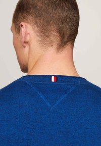 Blauer Pullover mit runden Ausschnitt und strukturiertem Stoff, der an dem hinteren Kragen mit einem kleinen Akzent in Rot, Weiß und Blau versehen ist.