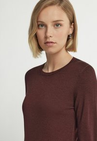 Burgundy långärmad stickad topp med rund halsringning, slät textur och rena sömmar, delvis synlig mot en neutral bakgrund.