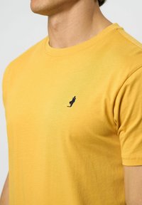 Tricou din bumbac galben, cu mâneci scurte, gât rotund și un mic detaliu brodat în albastru închis pe piept. Textură netedă și design slim.