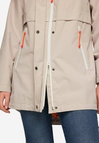 Ljusbeige vattentät jacka med vita dragkedjor och orange dragflikar. Den har två fickor med dragkedja och en tryckknappsstängning.