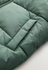 Tissu matelassé vert avec poche rectangulaire cousue et détail de rabat anglé sur un vêtement rembourré.
