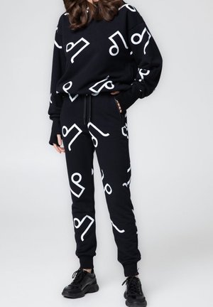 Osoba ubrana w czarną bluzę i spodnie dresowe z dużymi białymi abstrakcyjnymi wzorami, w połączeniu z czarnymi trampkami, stojąca na jednolitym tle.