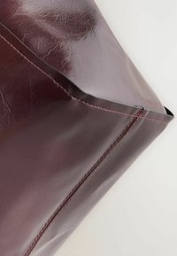 Bordeaux leren tas heeft een gladde textuur met zichtbare nerf. De randen zijn gestikt met contrasterend roze garen, wat een modern ontwerp verwezenlijkt.