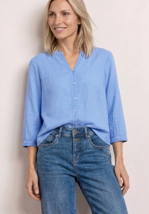 Femme aux cheveux blonds portant un chemisier bleu clair boutonné avec des manches 3/4 et un jean bleu taille haute, souriante et regardant vers la gauche.