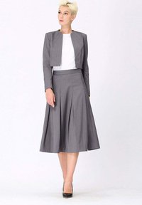 Femme avec les cheveux blonds coupés portant une veste grise courte, un haut blanc, une jupe midi plissée grise et des talons noirs, se tenant contre un fond blanc.