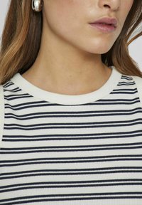 Stripet topp med horisontale marineblå striper på en hvit ribbestrikket stoff, med rund hals og tettsittende design. Sølvfargede smykkeører synlige.
