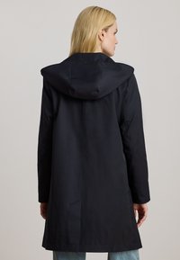 Gabardina negra larga con capucha, que presenta una textura suave y un diseño minimalista. La gabardina llega hasta la mitad del muslo y tiene un corte recto.