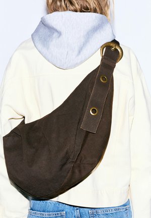 Osoba nosi svetlú mikinu s kapucňou a bielu bundu s veľkou tmavohnedou semišovou crossbody taškou s mosadznou prackou a očkami.