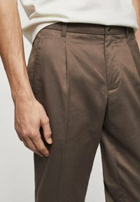Pantalones marrones a medida con una textura suave, que presentan pliegues laterales y un diseño de frente plano. La cinturilla incluye pasadores para cinturón y un cierre de botón.