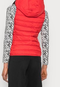Kobieta w czerwonej kurtce puchowej z kapturem, nosząca czarno-biały top w paski zebry z długim rękawem oraz czarne spodnie, pokazana z tyłu.