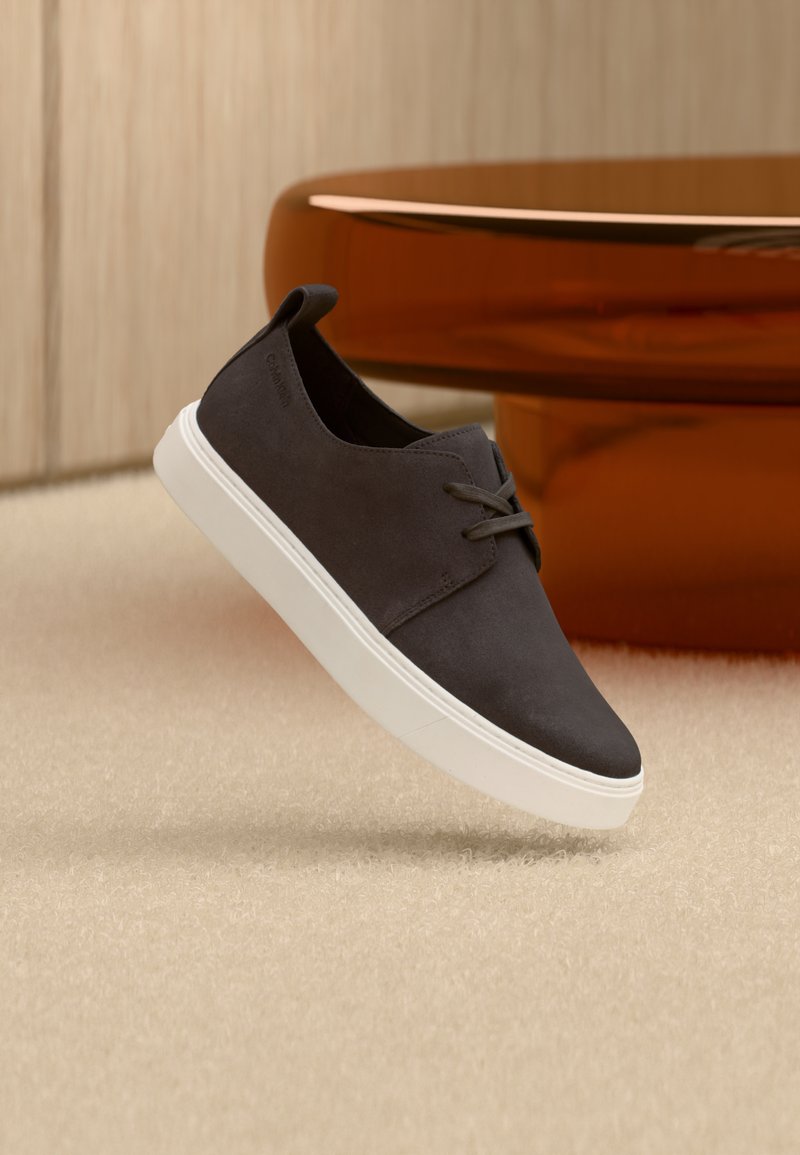 Σκούρο γκρι σουέτ casual παπούτσι με κορδόνια και λευκή σόλα, κρεμασμένο πάνω από μπεζ χαλί, με καφετί διαφανές μοντέρνο τραπέζι στο φόντο.