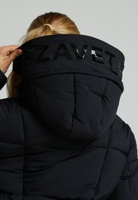 Crna izolirana jakna s visokim ovratnikom i kapuljačom. Ima sjajni "ZAVET" logo na leđima i strukturirani kviltani dizajn.