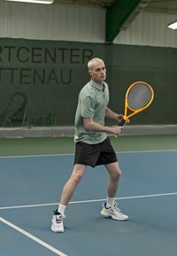 Mies tennispelaaja vaaleanvihreässä polo-paidassa ja mustissa shortseissa, pitelee oranssia mailaa sinisellä kentällä, taustalla verkko.