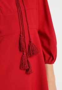 Červené šaty s hladkým materiálem, nabíranými rukávy a dlouhými stužkami s třásněmi. Významné detaily zahrnují texturované konce třásní a jednoduchý design.