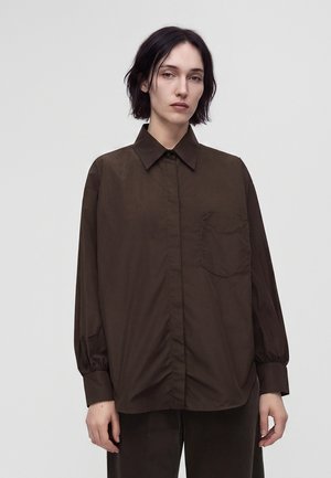 Femme aux cheveux courts et foncés portant une chemise marron foncé ample à boutons avec une poche avant et un pantalon foncé assorti sur un fond uni.