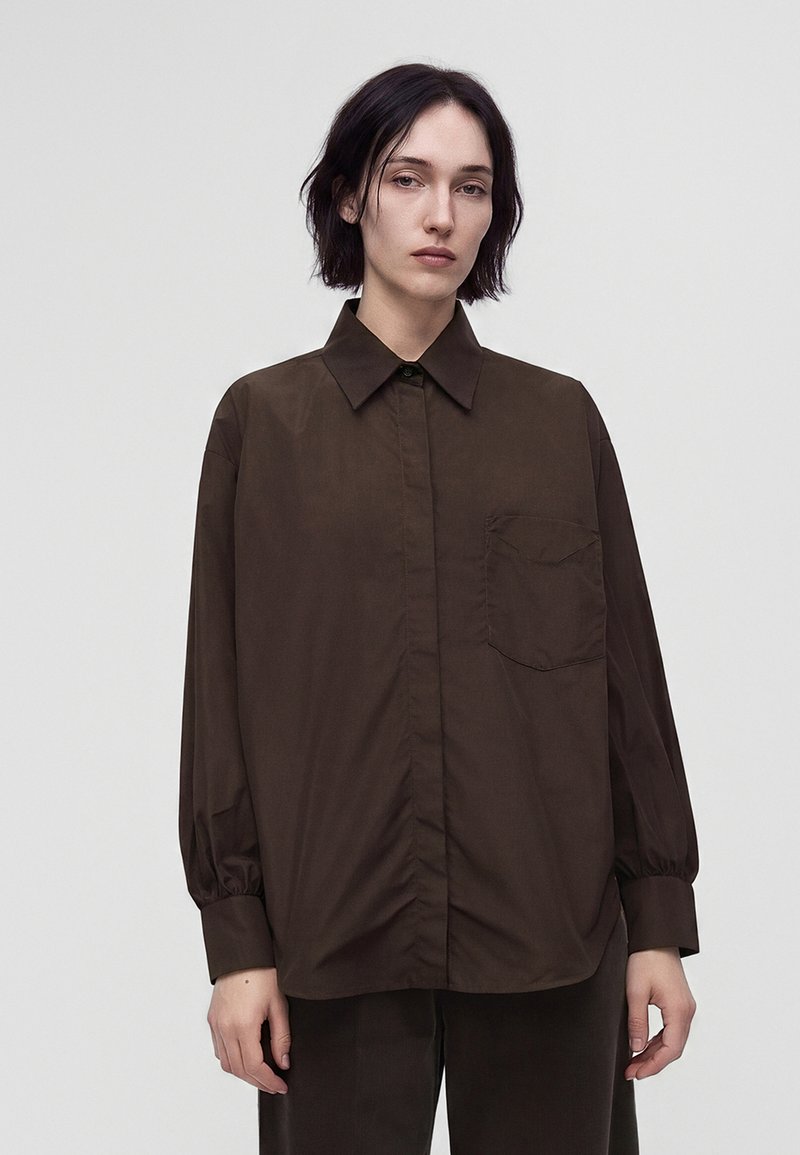 Femme aux cheveux courts et foncés portant une chemise marron foncé ample à boutons avec une poche avant et un pantalon foncé assorti sur un fond uni.