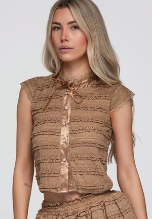 Femme aux longs cheveux blonds portant un haut marron en dentelle sans manches avec des boutons et une jupe froncée assortie, posant devant un fond uni.