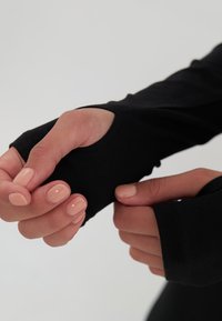 Crna majica s dugim rukavima i rupama za palčeve, mekana tkanina, prilagođeni dizajn. Ruke s neutralnim lakom za nokte spojene su blizu manžeta.
